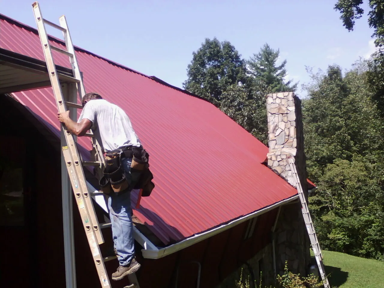Roofer on a ladder beside a red metal roof and stone chimney — Soffit & Fascia Repair by Roofing Brothers in Warfield